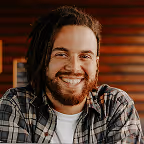 Smiling man with beard and long hair wearing a plaid shirt in a warmly lit room.